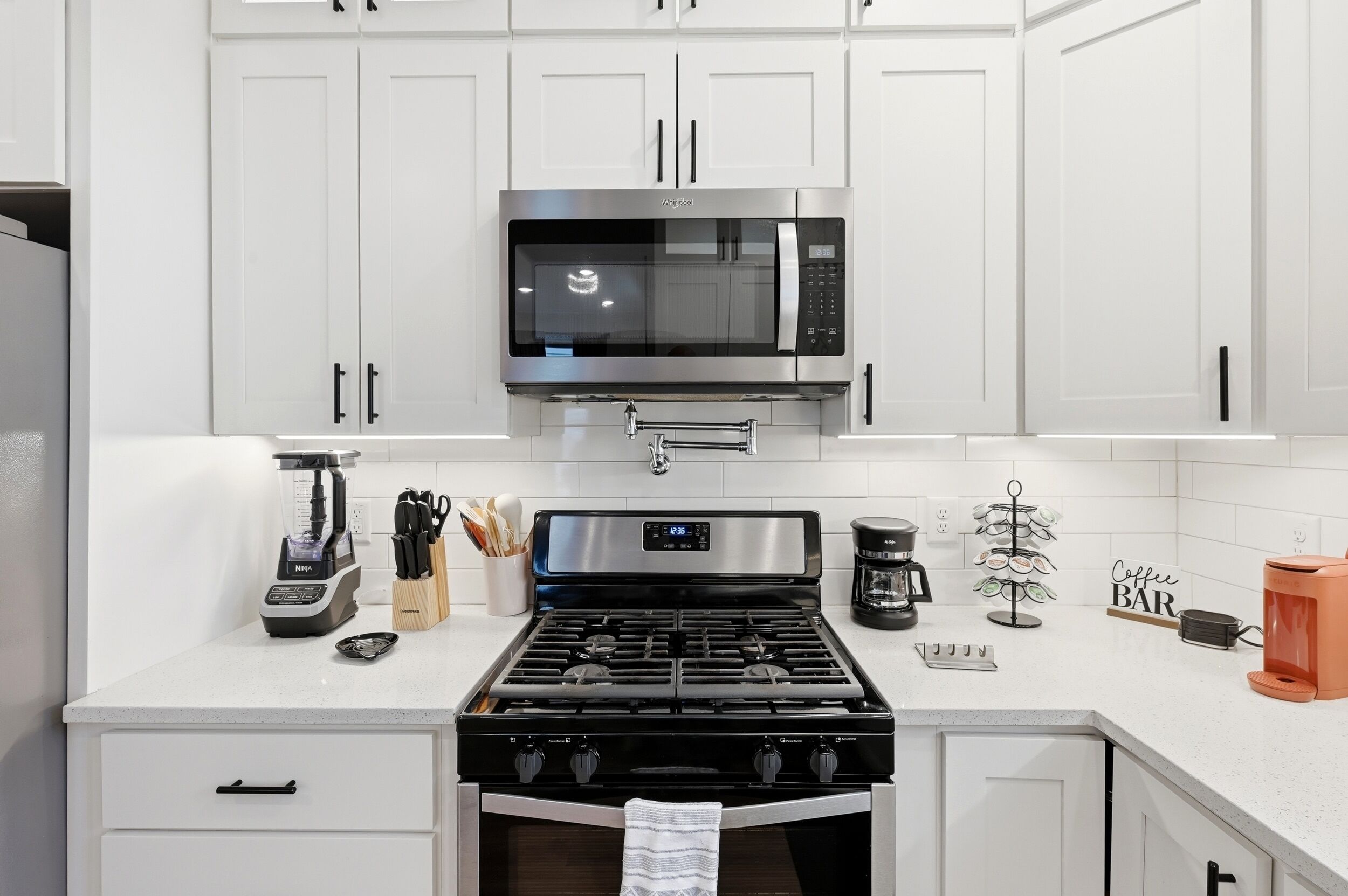 A modern kitchen with white cabinets, a stainless steel microwave, a gas stove, a coffee maker, and a coffee bar sign.