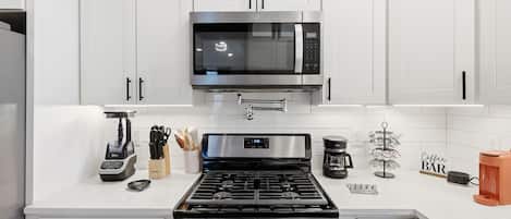 A modern kitchen with white cabinets, a stainless steel microwave, a gas stove, a coffee maker, and a coffee bar sign.