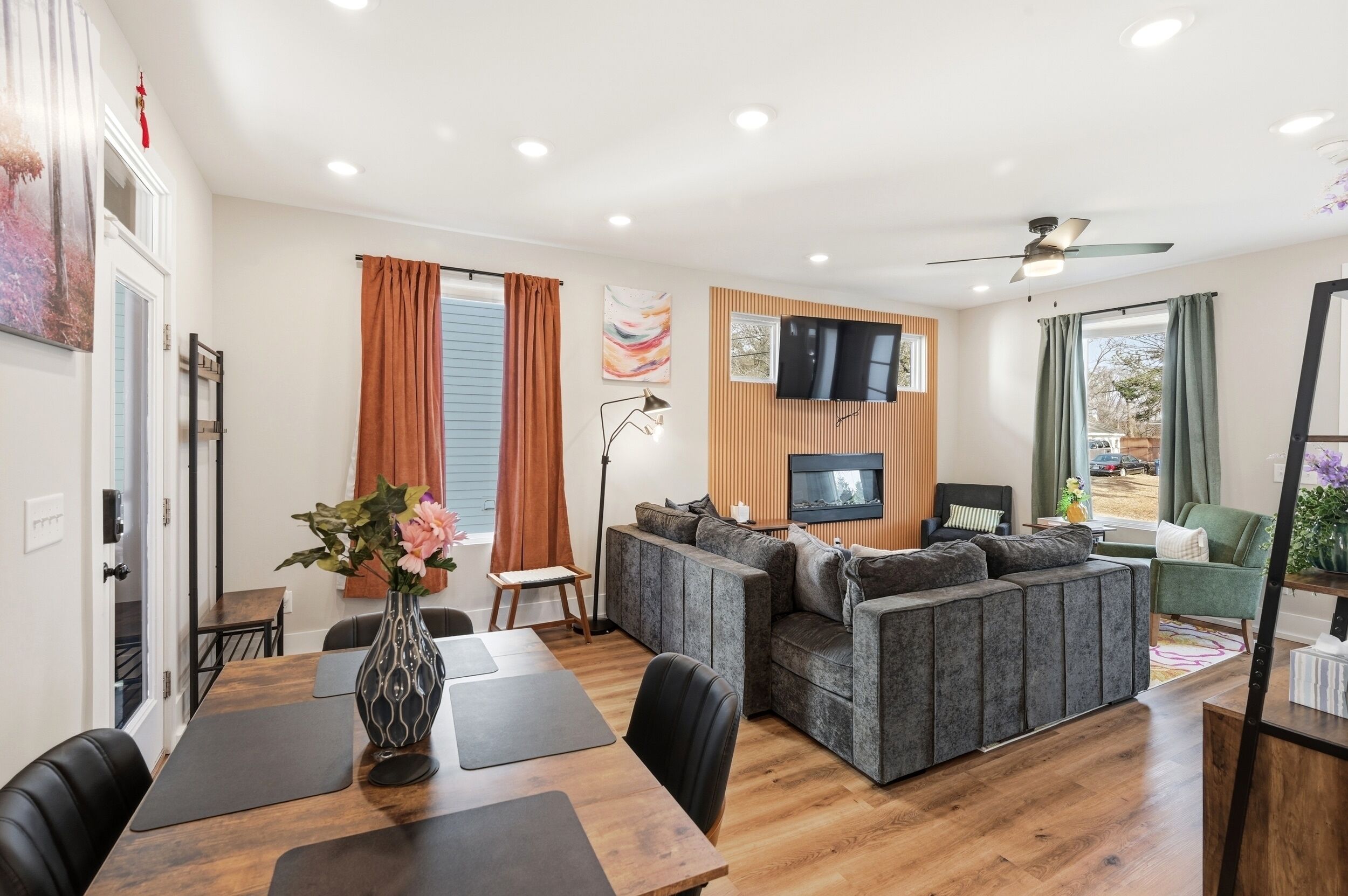 A modern living room with a sofa, a dining table with chairs, a TV mounted on the wall, and a ceiling fan.