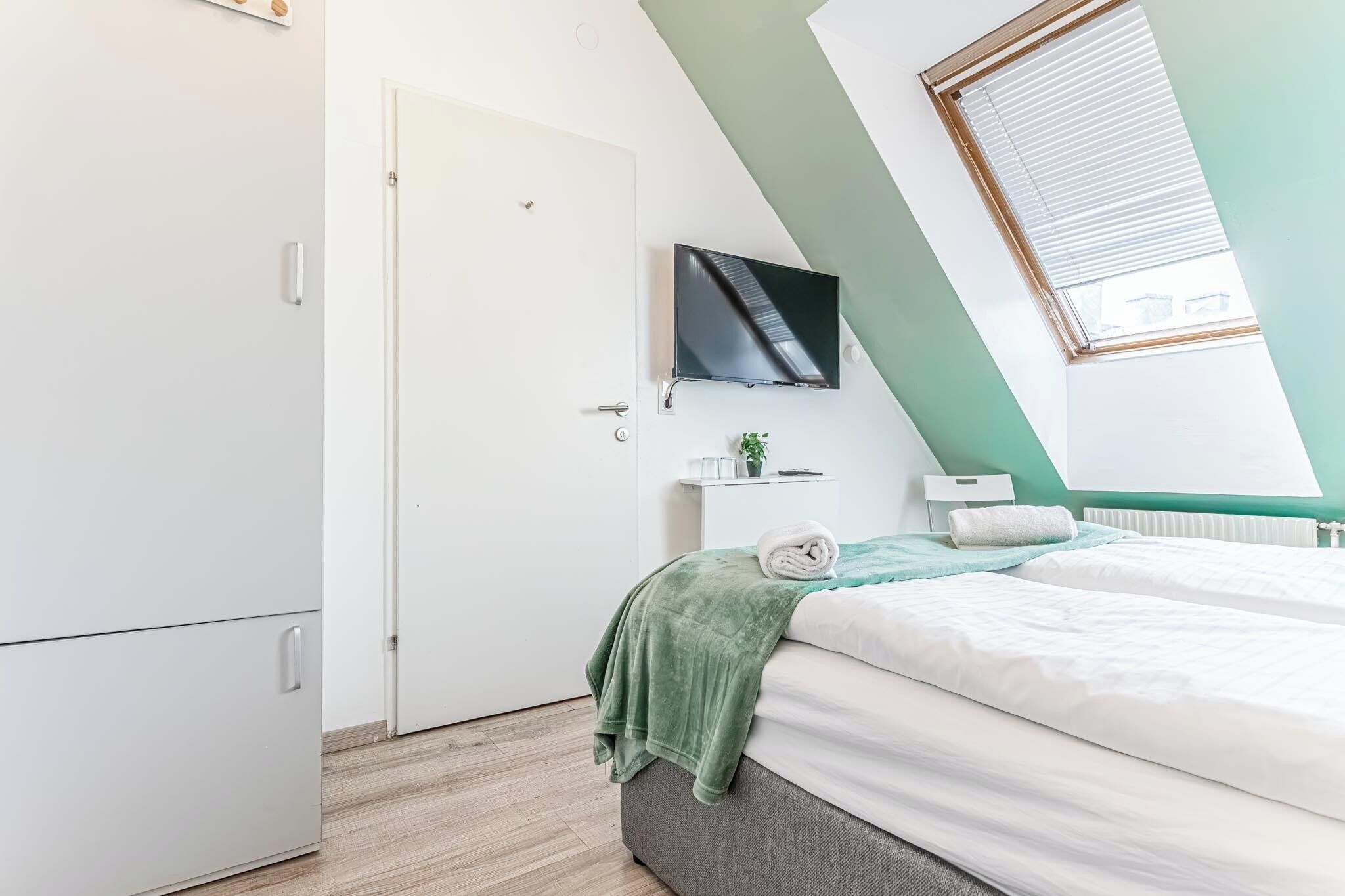 A bedroom with a bed, a TV mounted on the wall, a door, and a window with blinds.