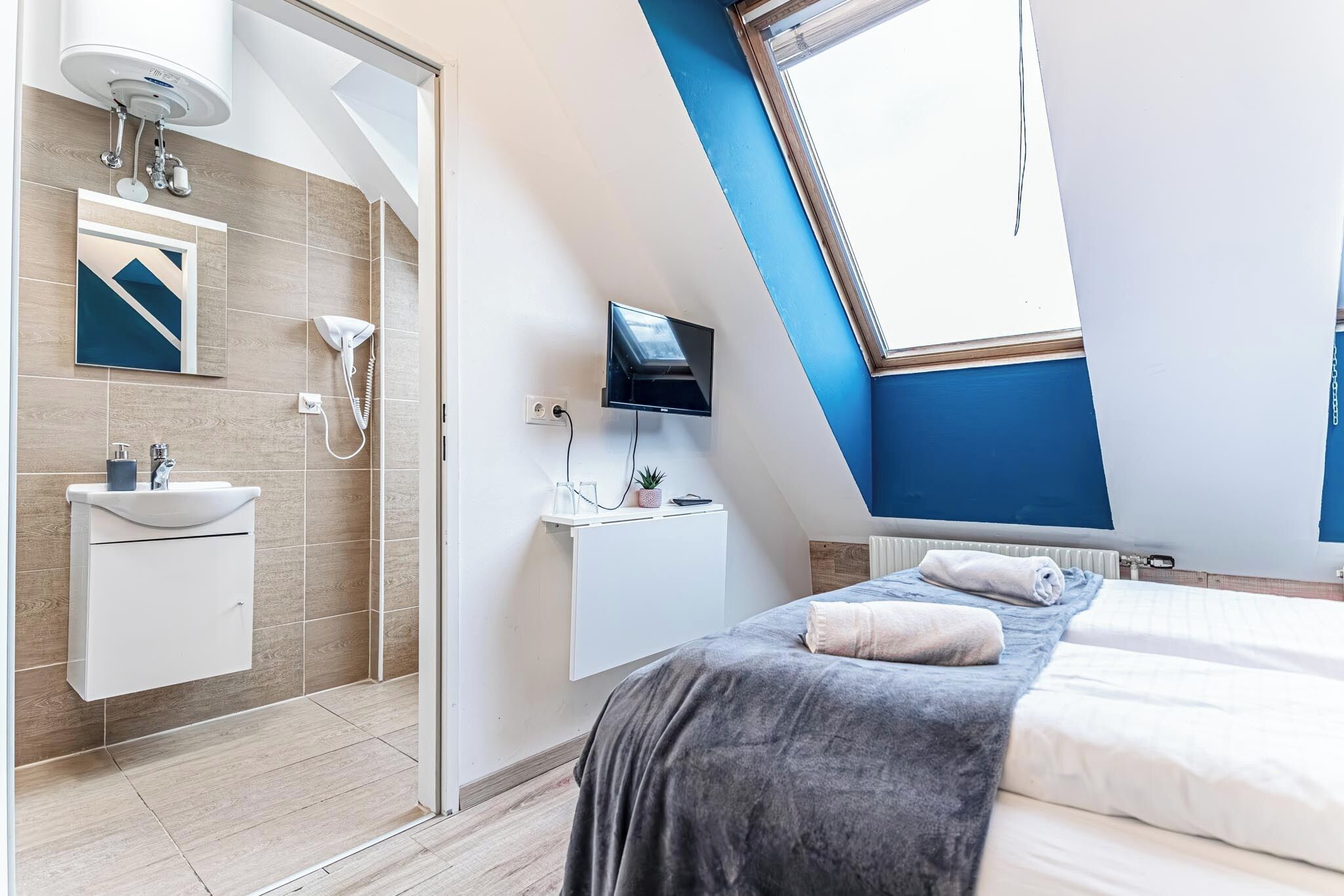 Une chambre moderne avec un puits de lumière, un téléviseur fixé au mur, une table de chevet agrémentée d’une plante et un grand lit recouvert d’une couverture.