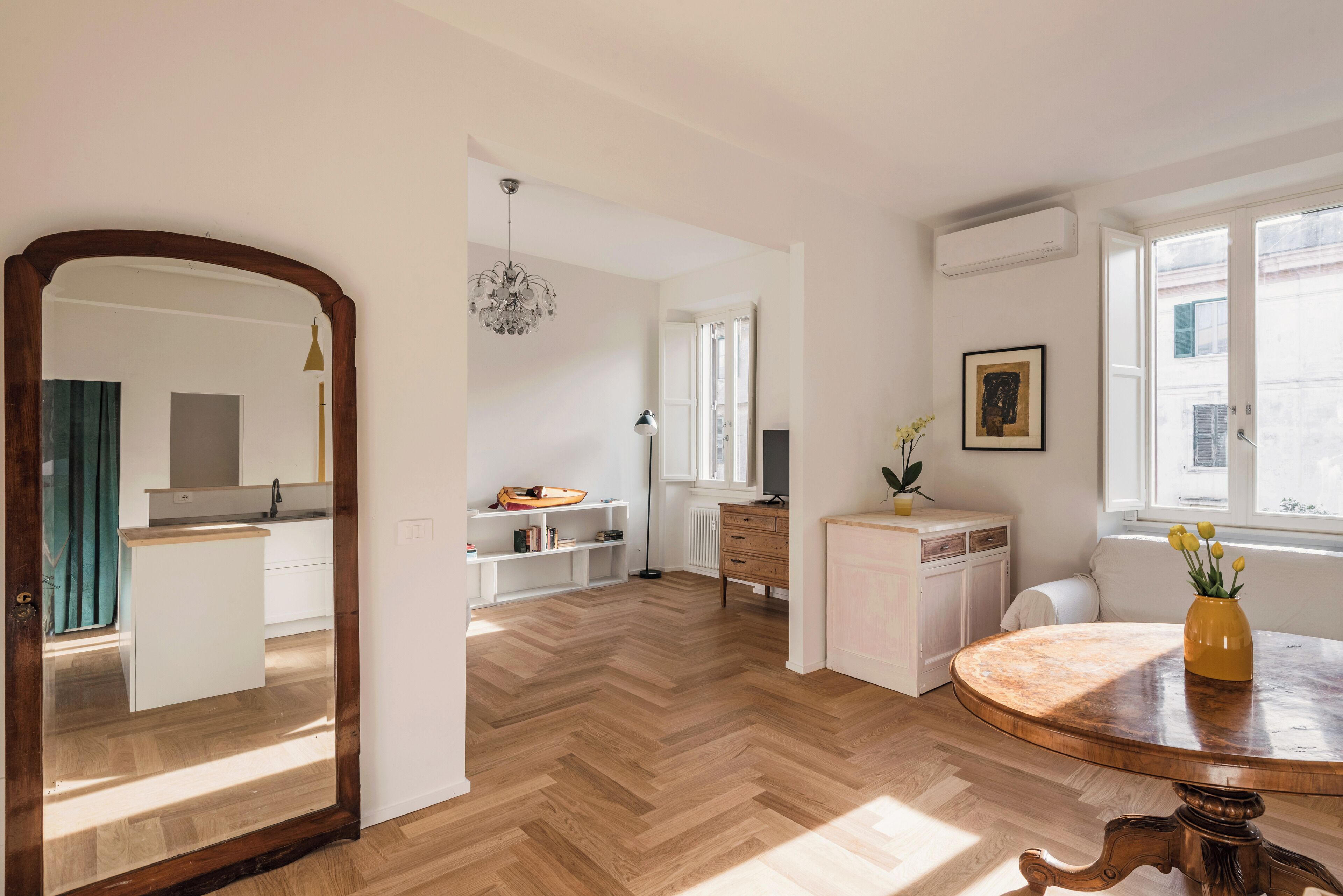 Una cucina moderna con mobili bianchi, pavimento in legno a spina di pesce e un tavolo rotondo in legno con un vaso di fiori.
