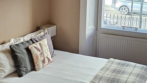 Un dormitorio con cama, ventana con persianas, araña y un cuadro en la pared.