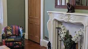 Una habitación con piso de madera, una mesa de madera, una silla con estampado floral colorido, una chimenea con repisa blanca y un candelabro.