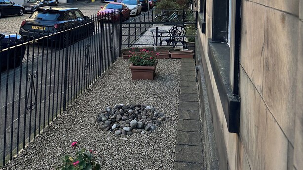 Una estrecha calle urbana con automóviles estacionados, un muro de piedra y un edificio de fachada pétrea.