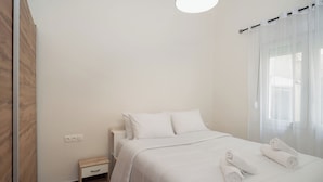 Un lit bien fait, avec du linge de lit blanc et des serviettes pliées, une table de chevet et une fenêtre ornée de rideaux légers.