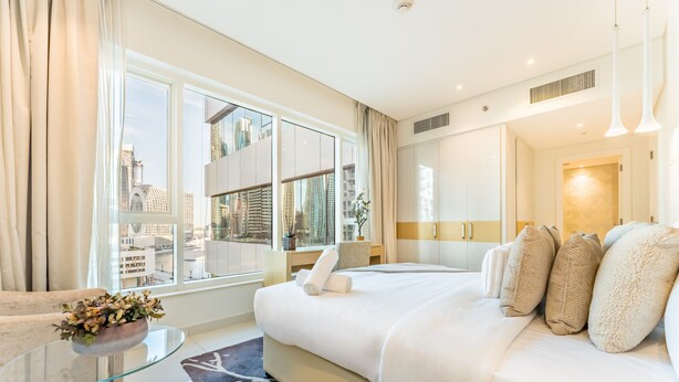 Une chambre d’hôtel moderne, dotée d’un grand lit, d’une table basse en verre et offrant une vue sur le paysage urbain grâce à de grandes fenêtres.