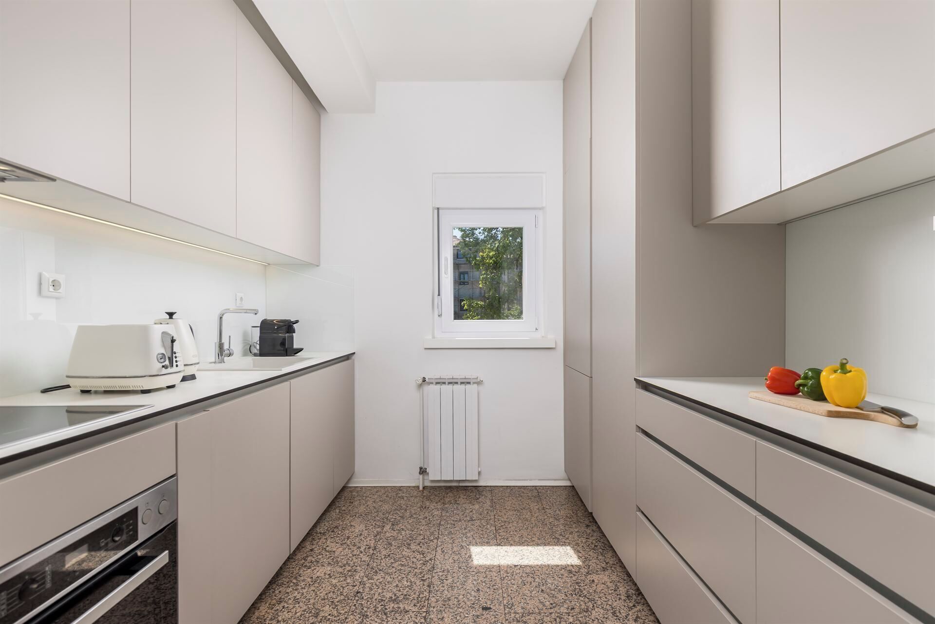 Una cocina estrecha con armarios blancos, horno empotrado, cafetera y ventana con vistas al verdor.