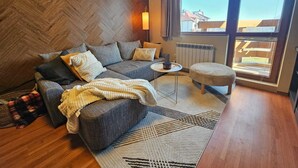 Ein modernes Wohnzimmer mit Fischgrätparkett, einer grauen Sofagarnitur, einem weißen Teppich und Blick auf Gebäude durch große Fenster.