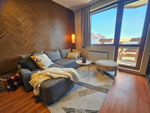 Ein modernes Wohnzimmer mit Fischgrätparkett, einer grauen Sofagarnitur, einem weißen Teppich und Blick auf Gebäude durch große Fenster.