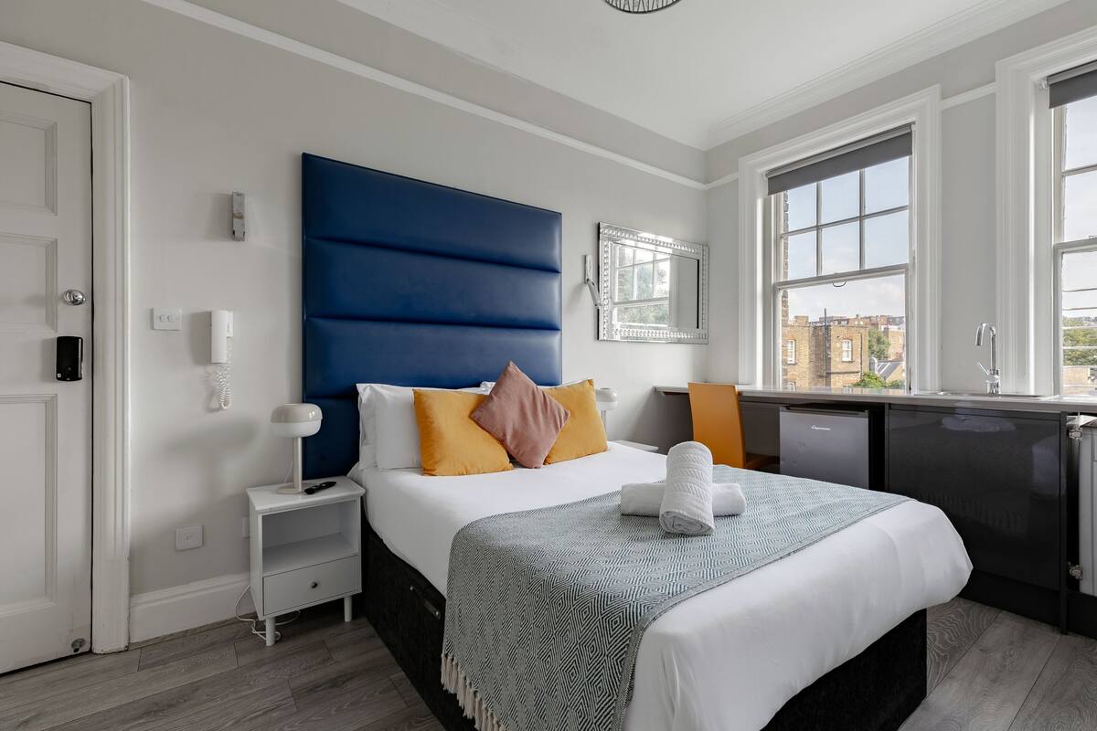 A modern hotel room with a large bed, a bedside table, a desk, and a view of the city.