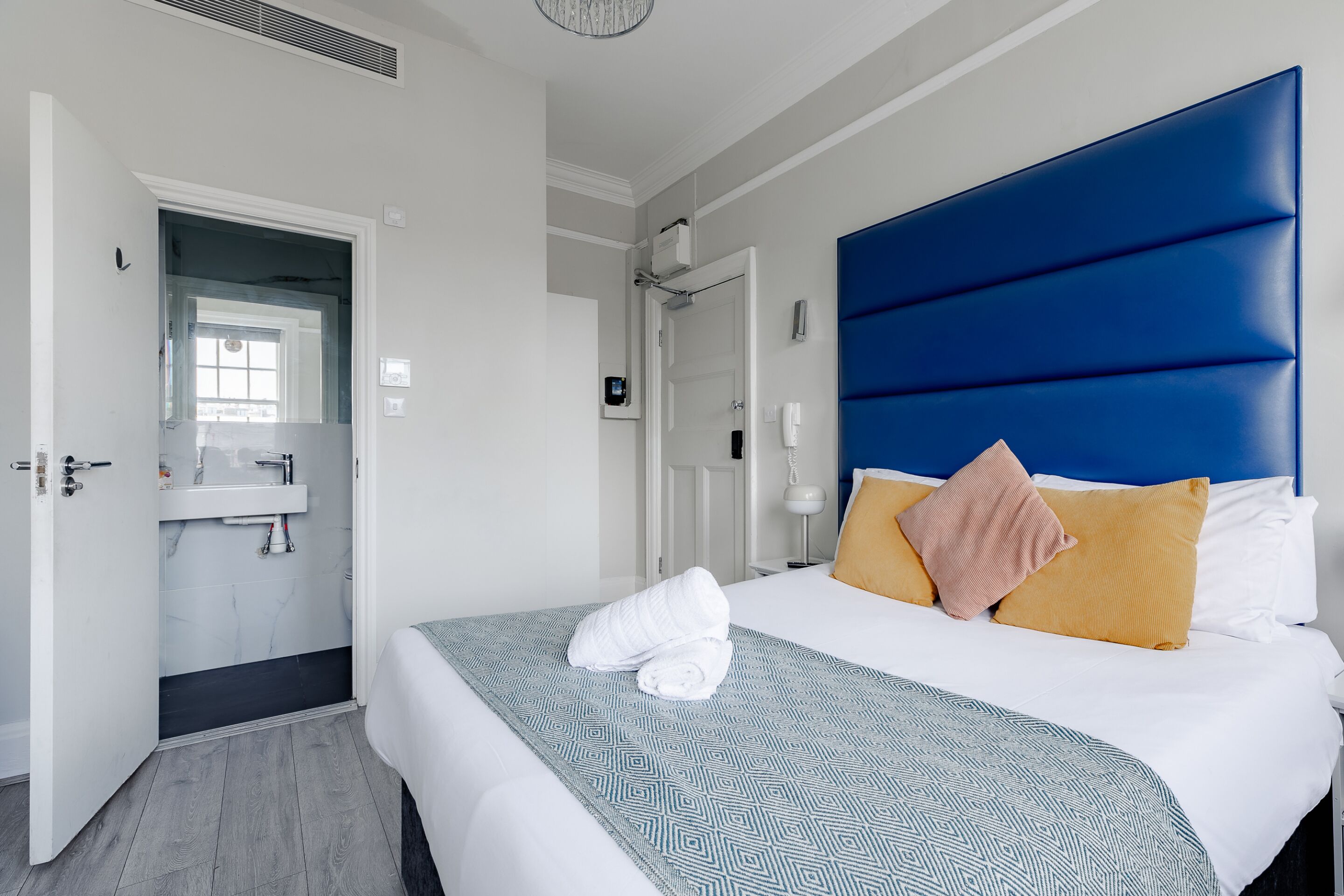 Ett hotellrum med en stor säng, en blå sänggavel, ett vitt nattduksbord och ett badrum som syns genom en öppen dörr.