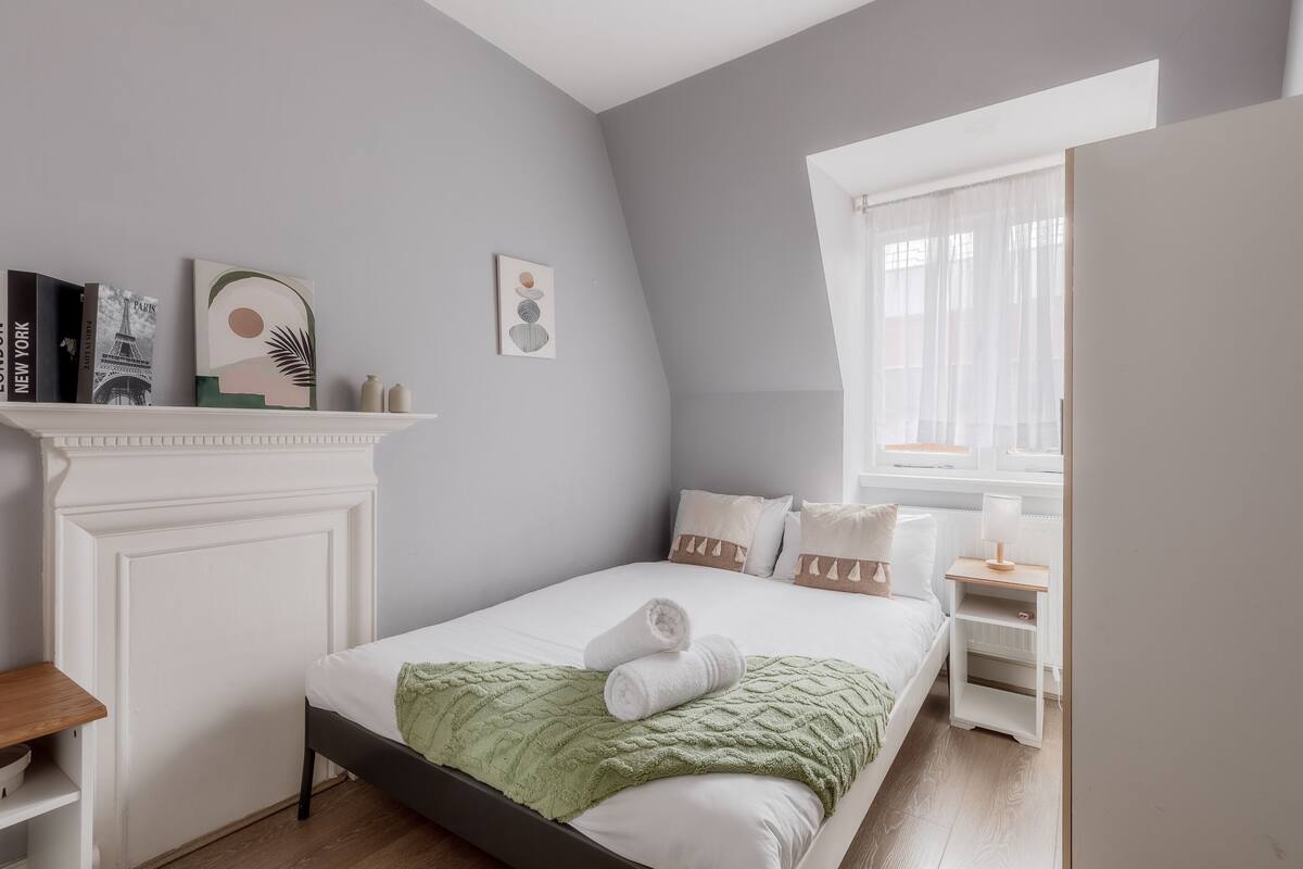 A neatly arranged bedroom with a bed, bedside tables, a nightstand, a fireplace, and wall art.