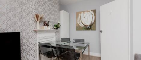 A modern dining area with a glass table, four chairs, a TV, a fireplace, and a painting on the wall.