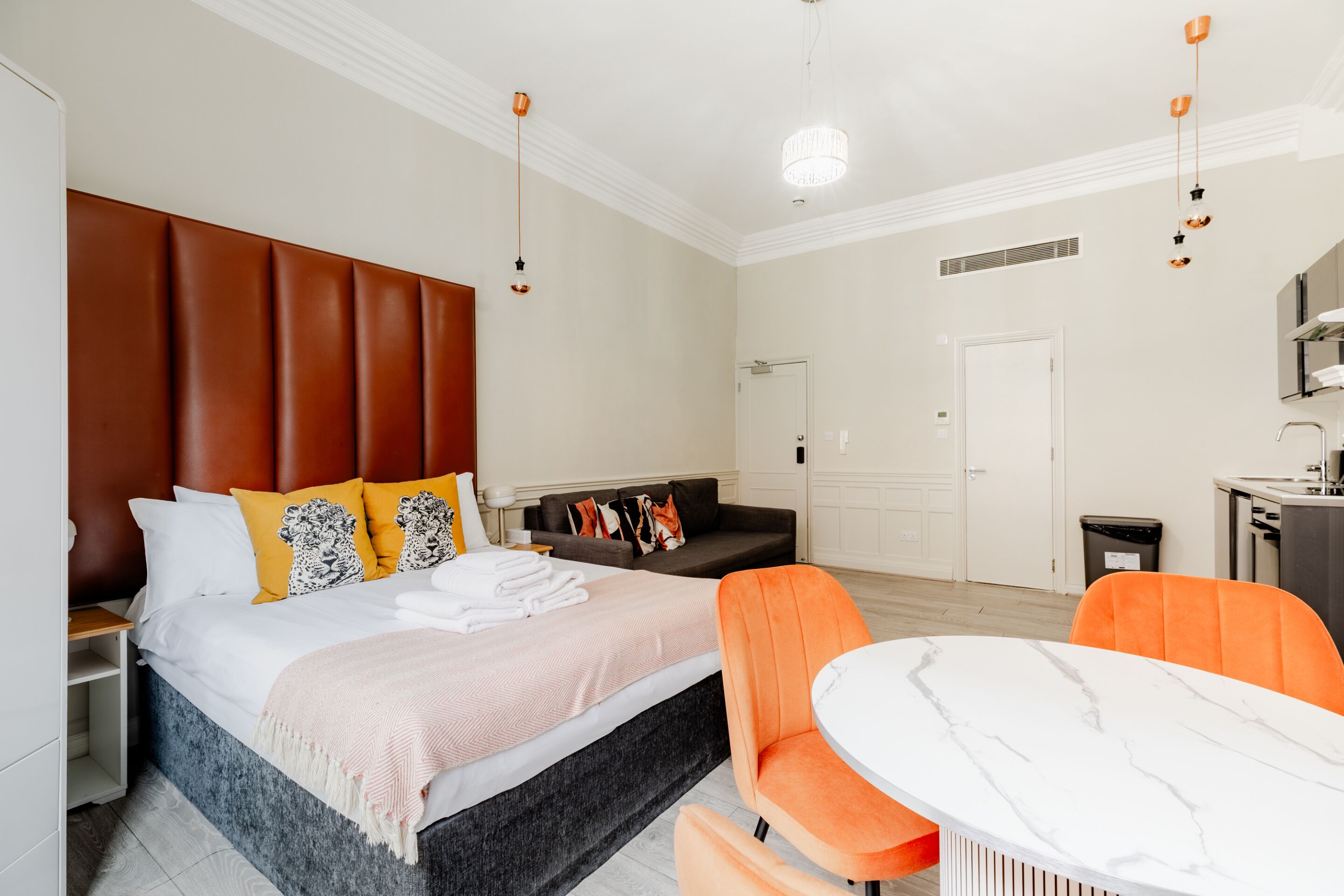 A modern hotel room with a large bed, a sofa, a dining table, and a kitchenette.