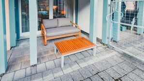 A wooden bench and a grey sofa on a paved patio with a glass door and a metal gate. - Surfers Deck | Terrace & Lake View (Därligen)