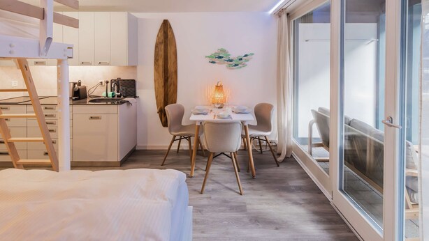 A compact hotel room with a bed, a dining area with chairs and a table, a surfboard, and a large window. - Surfers Deck | Terrace & Lake View (Därligen)