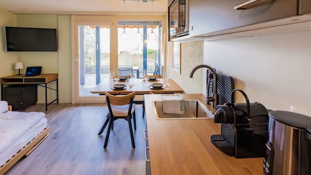 Una habitación compacta con cama, escritorio, mesa de comedor con sillas, kitchenette con cafetera y televisor.