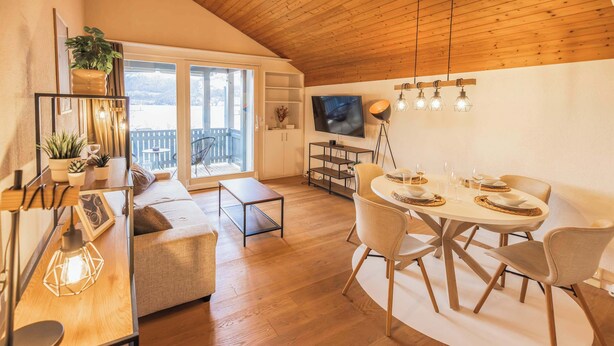A modern living room with a wooden ceiling, a dining area with a round table and chairs, a sofa, a TV, and a balcony with a view. - Surfers Lodge | Lake View & Pool (Därligen)