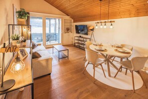 Una sala de estar moderna con techo de madera, un comedor con mesa y sillas redondas, un sofá, un televisor y un balcón con vistas.