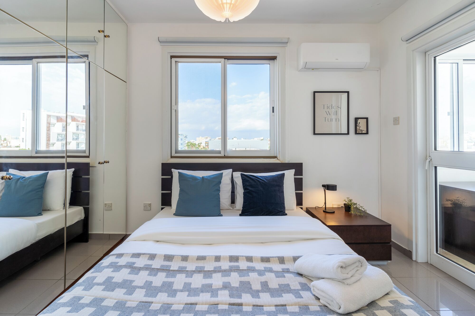 A modern bedroom with a large bed, two windows, a bedside table with a lamp, and a view of the city.