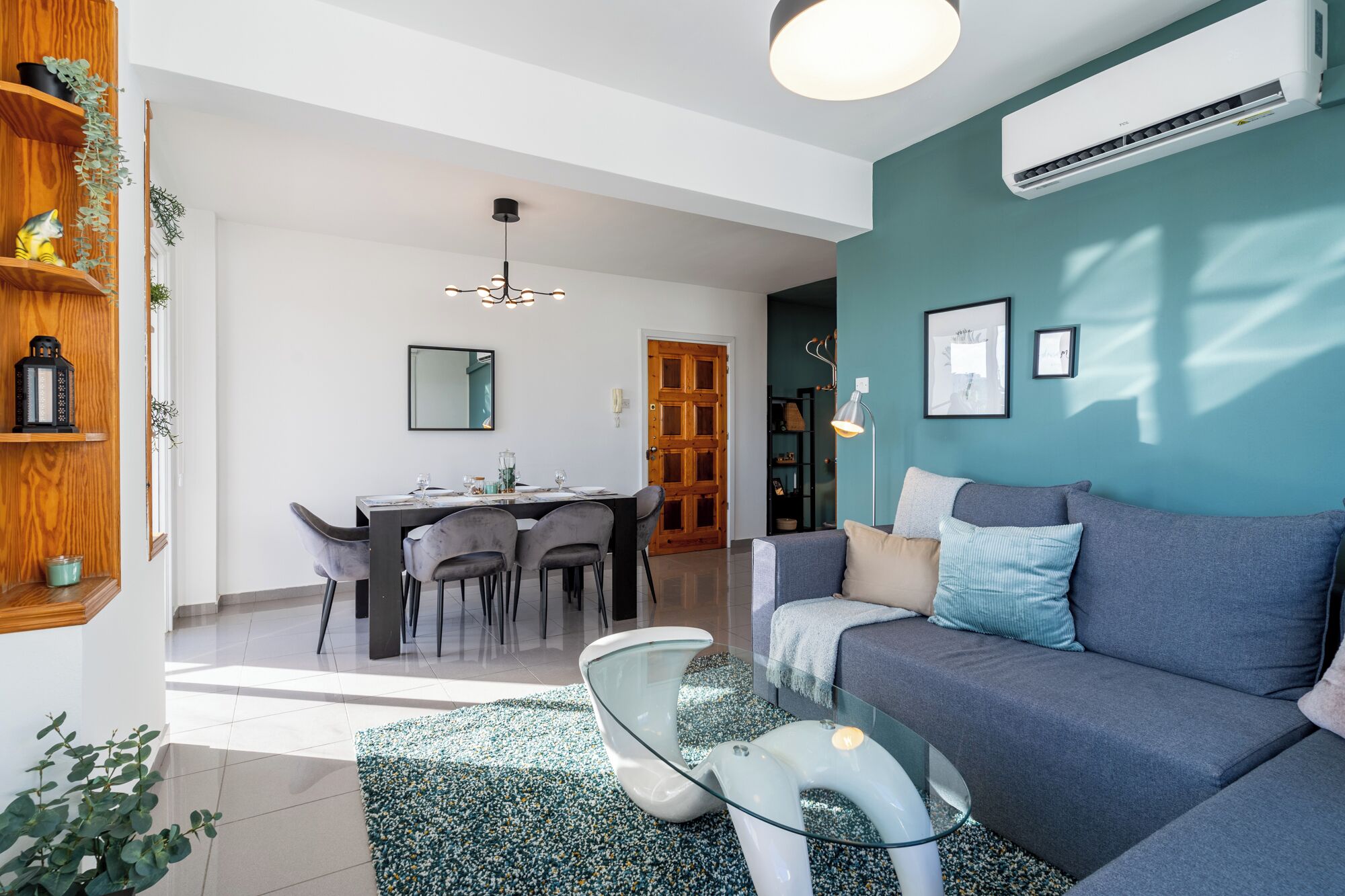 Una sala de estar moderna con una pared azul, un sofá gris, una mesa de centro de vidrio y un comedor con puerta de madera.