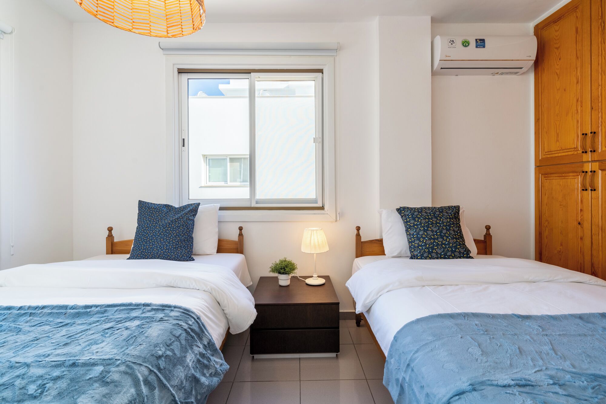 A bedroom with two beds, a window, an air conditioner, and a bedside table with a lamp.