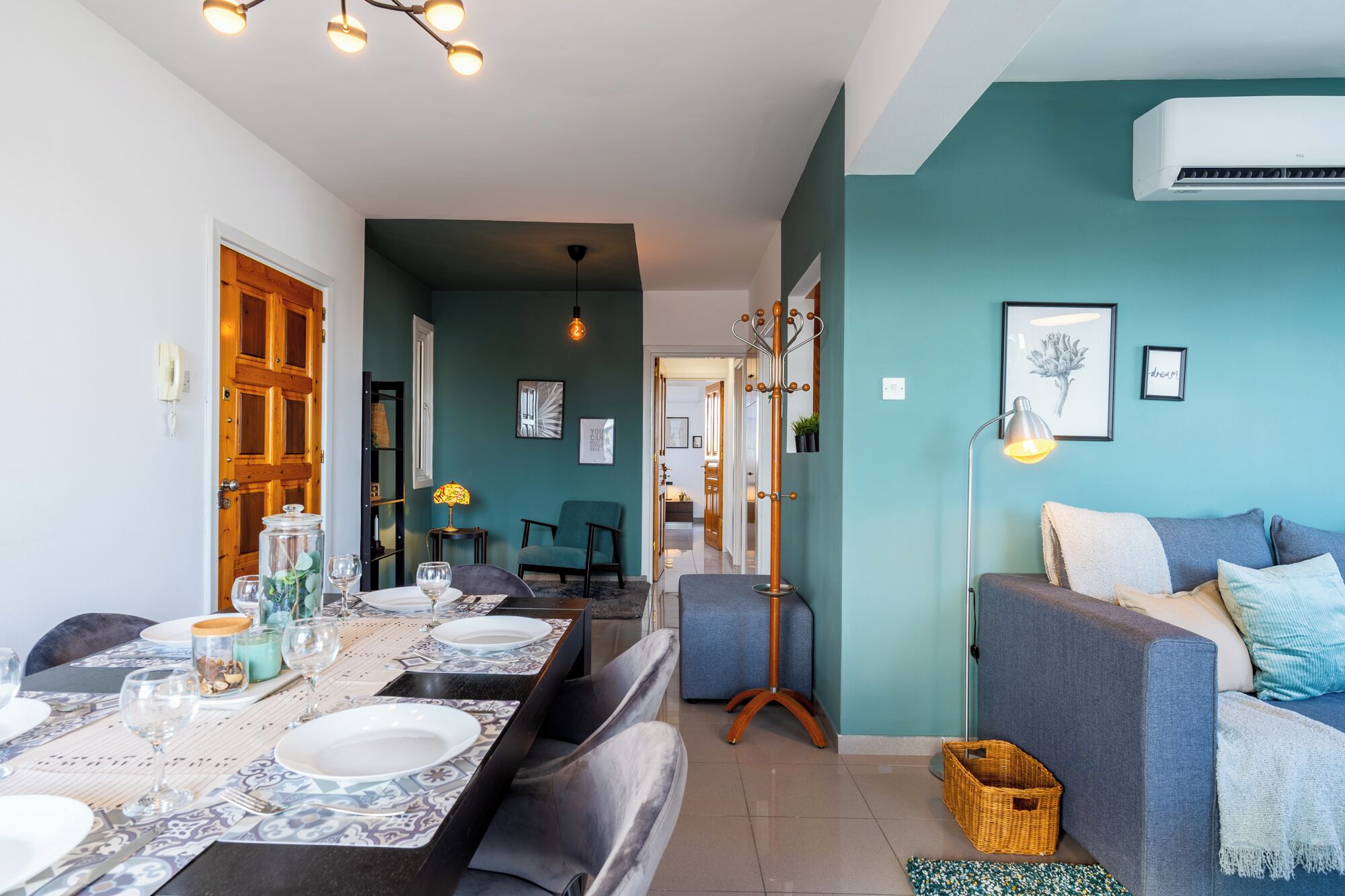 Un espace repas moderne avec une longue table dressée pour quatre personnes, un canapé bleu et un panier en osier posé sur le sol.