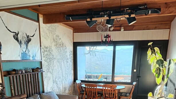 Ein gemütliches Wohnzimmer mit Holzboden, einer Couch, einem Esstisch mit Stühlen und einem großen Fenster mit Blick ins Freie.