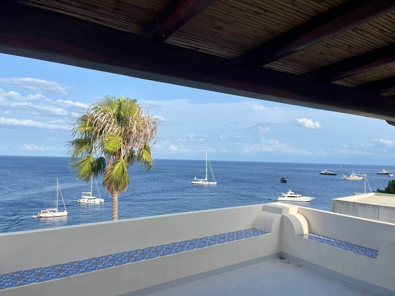 A balcony with a view of the sea and boats, a palm tree, and a tiled floor.