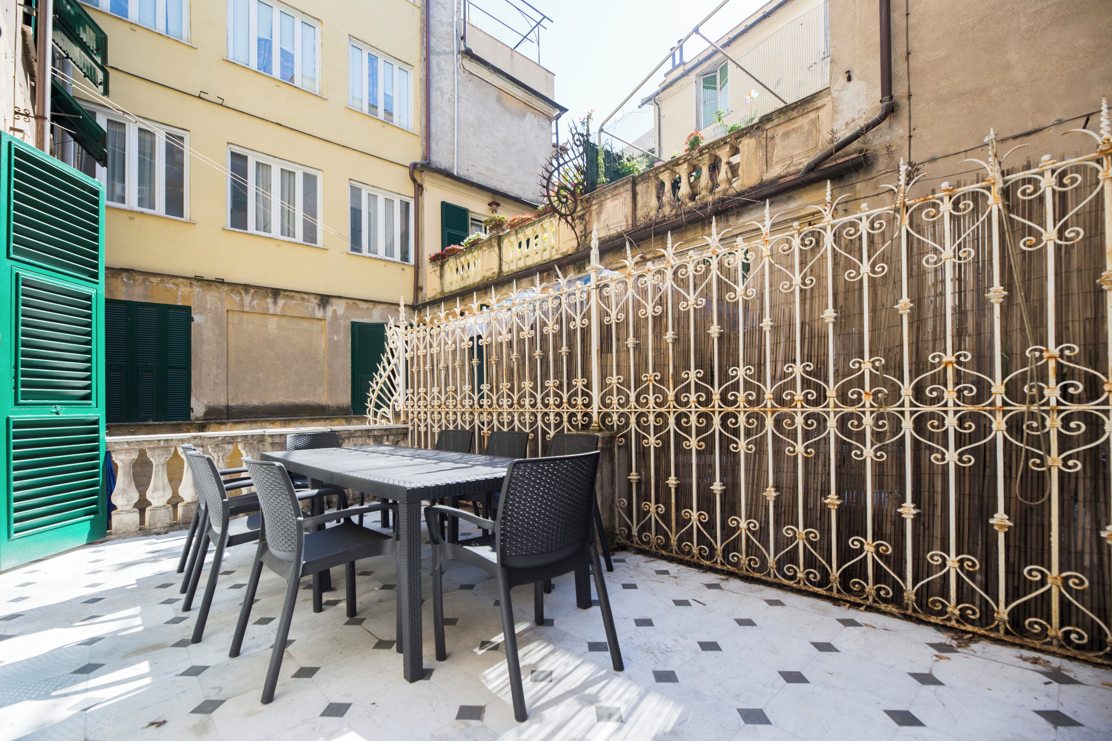 An outdoor patio with a table and chairs, a decorative metal fence, and a building with balconies and windows.