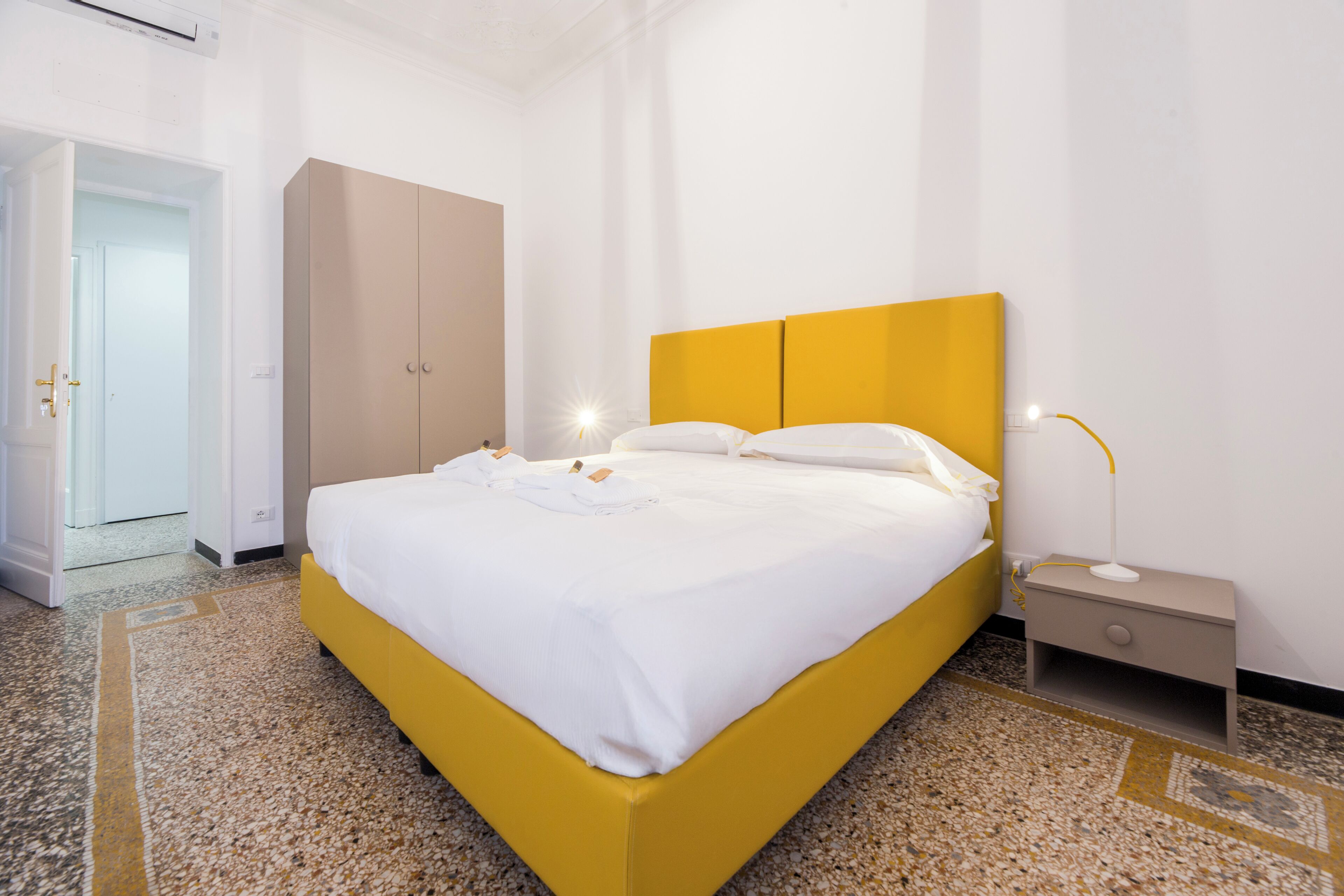A hotel room with a yellow bed, white bedding, a nightstand with a lamp, and a wardrobe.