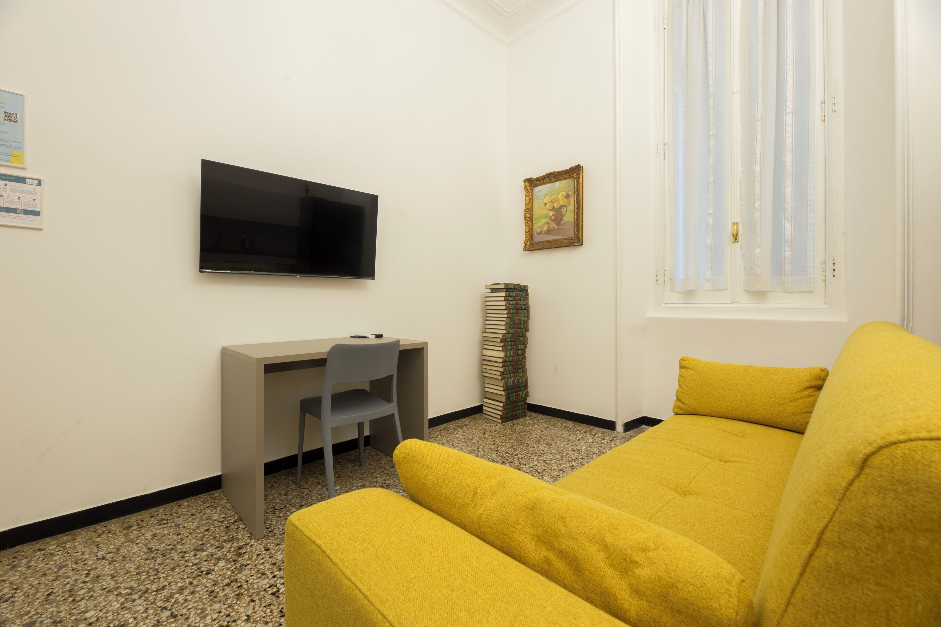 A room with a bed, a desk with a chair, a television, a window with curtains, and a framed picture on the wall.