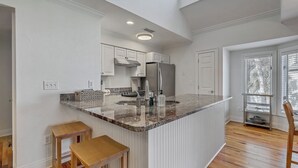 Eine Küche mit weißen Schränken, einer Granitarbeitsplatte und einem Edelstahlkühlschrank. Zwei Holzstühle und ein Fenster mit Jalousien sind ebenfalls vorhanden.