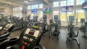 Ein Fitnessstudio mit mehreren Laufbändern und Crosstrainern, großen Fenstern und Bildschirmen, die Sportübertragungen zeigen.