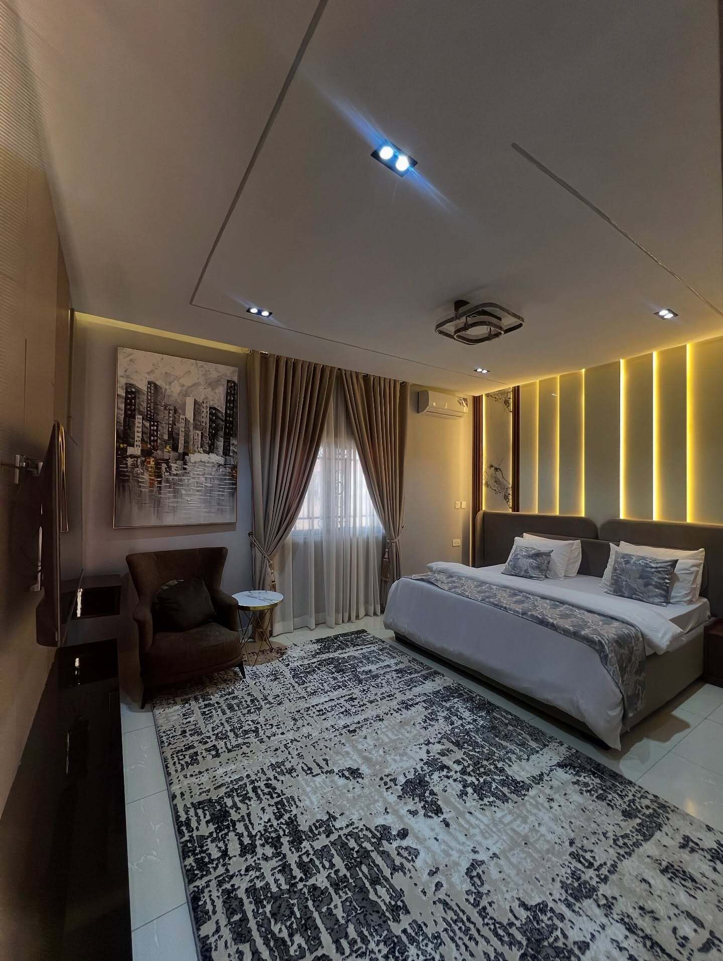 A modern bedroom with a large bed, a small round table, a dark armchair, and a cityscape painting on the wall.