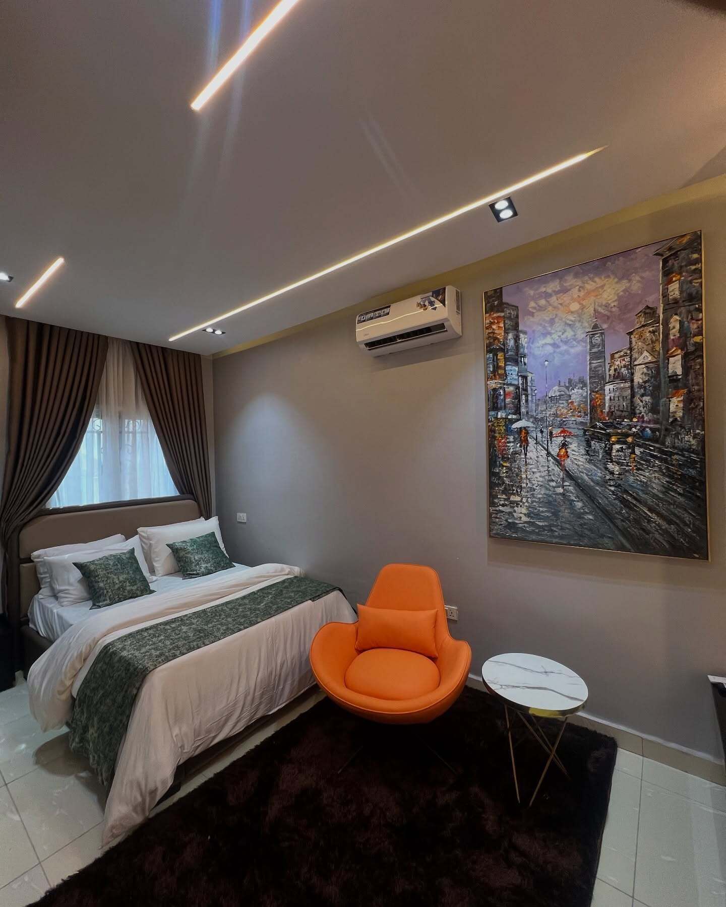 Un dormitorio moderno con cama, aire acondicionado, un cuadro, una silla y una mesita.