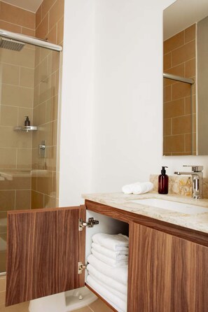 A bathroom with a shower, a sink with a marble countertop, a wooden cabinet with towels, and a mirror above the sink. - HOM Mineria Expanse Modern 2 BR in Mexico (Mexico City)