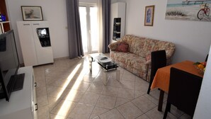 Uma sala de estar com sofá, mesa de centro, mesa de jantar com cadeiras, televisão e frigorífico.