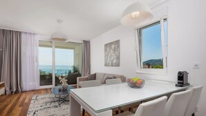 Ein modernes Wohnzimmer mit einem Glastisch, weißen Stühlen, einer Couch und Meerblick durch große Fenster.