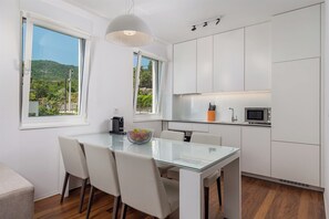 A modern kitchen with white cabinets, a glass dining table, and a view of greenery outside.