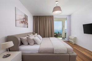 A modern hotel room with a large bed, a bedside table with a lamp, a flat-screen TV, and a view of the sea through a sliding glass door.