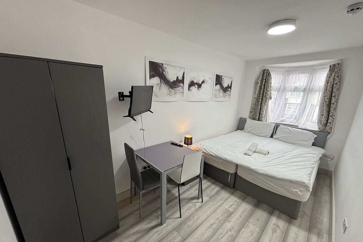 A compact hotel room with a bed, a desk with a chair, a wardrobe, and artwork on the wall.