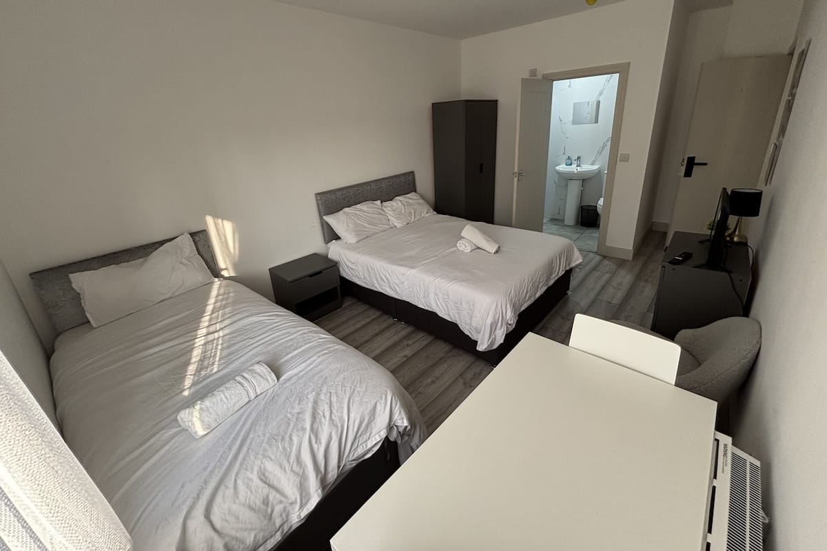 A hotel room with two beds, a small table, a desk, a mirror, and a bathroom visible through an open door.