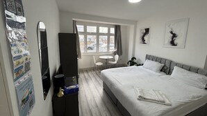 Quarto de hotel com cama, escrivaninha, cadeira, mesa pequena, espelho e janela com cortinas.