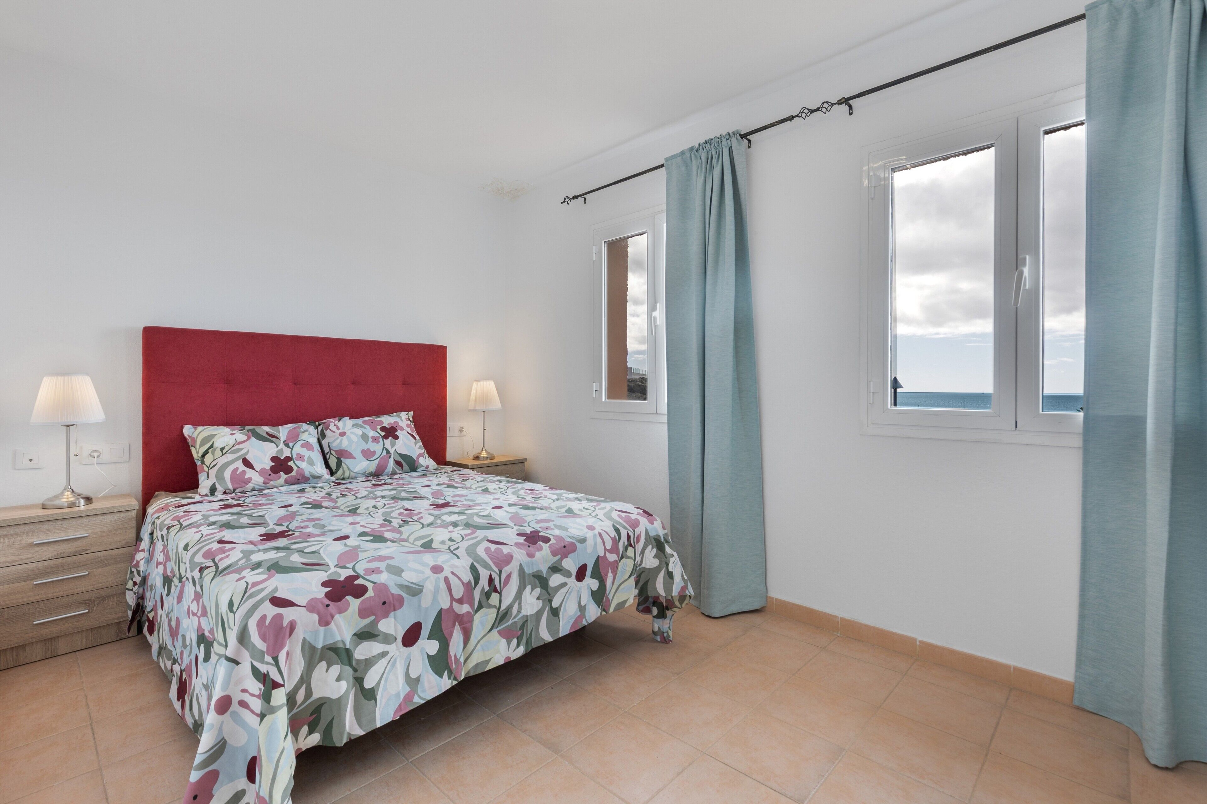 A bedroom with a red headboard, floral bedspread, two bedside lamps, a window with curtains, and a view of the sky.