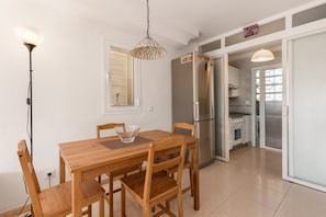 Ein Essbereich mit Holztisch und -stühlen, einer Pendelleuchte, einem Kühlschrank und einer durchgehenden Tür erkennbaren Küche.