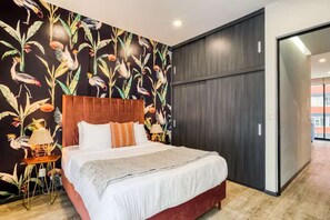 A bedroom with a large bed, a wooden headboard, a wall with a tropical bird and plant wallpaper, a small side table with a lamp, and a sliding door to the right. - HOM Roma Double l Spacious 2 Bedroom in Mexico City (Mexico City)
