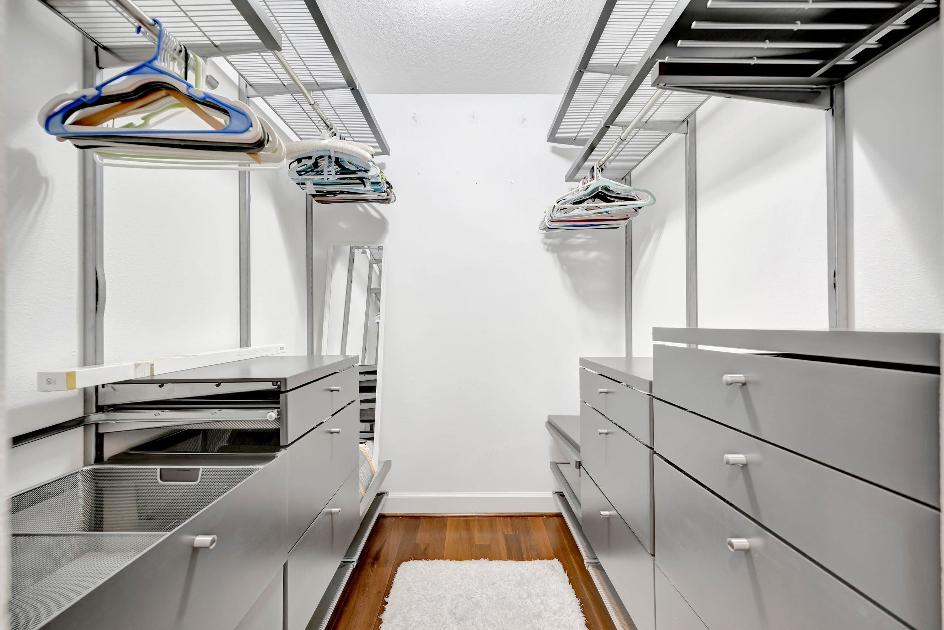 Un petit dressing étroit avec des étagères en métal, un tapis blanc et des vêtements suspendus.