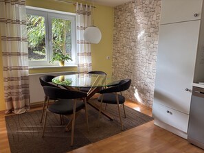 A dining area with a glass table and chairs, a window with curtains, a stone wall, and a white refrigerator. - Apartment 'Findus' with Private Terrace and Wi-Fi (Kempten-Haldenwang)
